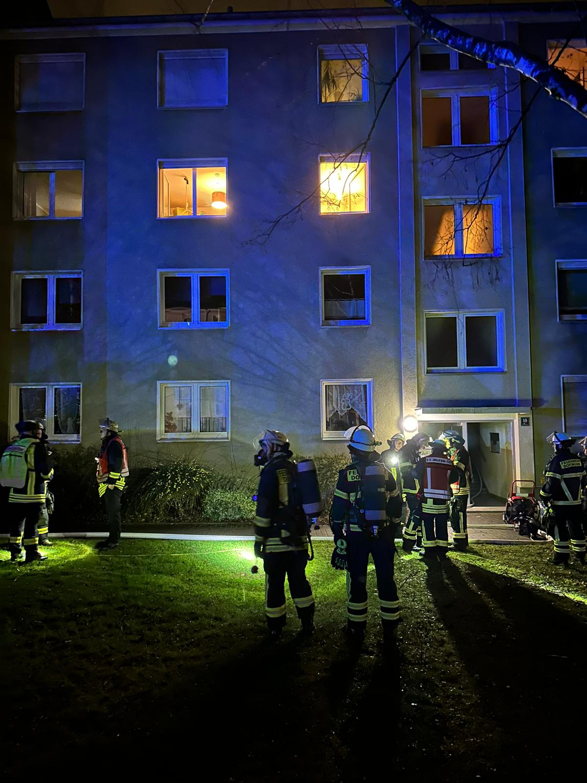 Feuerwehrangehörige stehen vor einem beleuchteten Mehrfamilienhaus in Bereitschaft